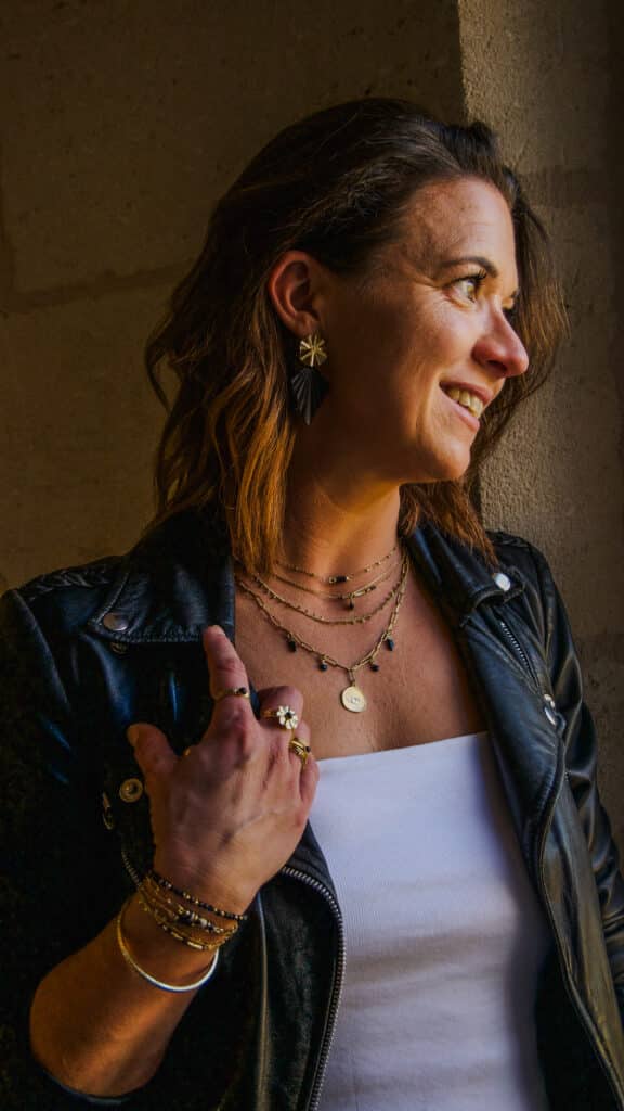 Femme souriante avec veste en cuir noir et débardeur blanc, portant des bijoux dorés superposés, y compris des colliers avec pendentif œil et des bagues.
