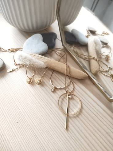 Collier long et boucles d'oreilles en or, agrémentés de perles, près de pierres Gua Sha cœur et d'un miroir sur bois clair.