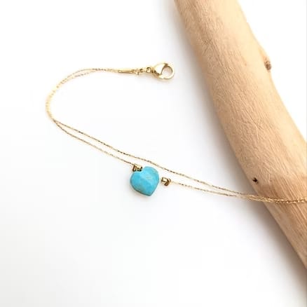 Bracelet double chaîne or fin avec cœur en turquoise facettée. Fermoir mousqueton doré visible, posé sur fond blanc et bois.