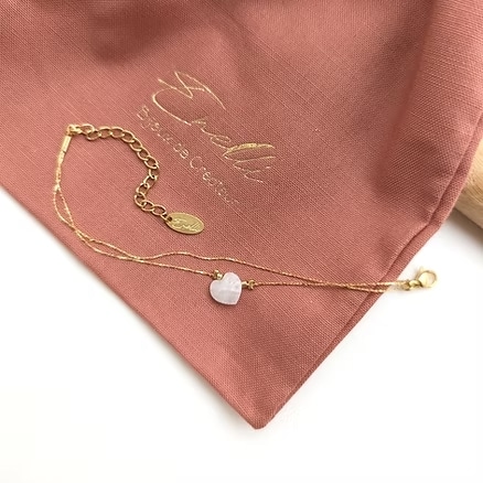 Délicat bracelet double chaîne doré, orné d'un pendentif cœur en quartz rose, posé sur un pochon rose brodé "Ebelli Bijoux de Créateur".