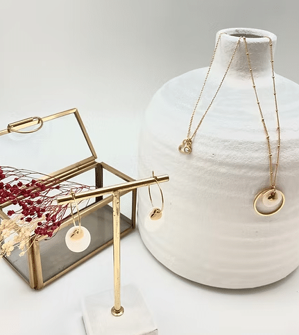 Bijoux dorés élégants. Boucles d'oreilles créoles et deux colliers pendants blancs et or, présentés devant un vase blanc texturé et une boîte en laiton.