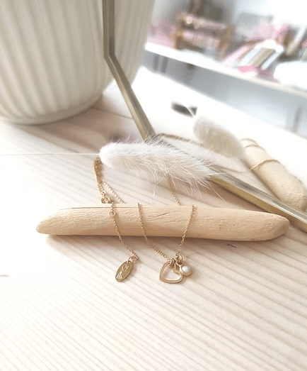 Collier doré fin, pendentif cœur ajouré et perle, présenté sur bois clair. Ambiance douce et lumineuse.