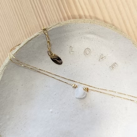 Collier fin en or avec pendentif cœur opalescent sur plateau céramique. La breloque porte la marque "Frolli". Le mot "LOVE" est gravé.