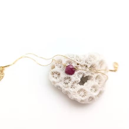 Collier fin en or avec pendentif rubis rouge brut posé sur un fragment de corail blanc texturé, fond blanc lumineux.