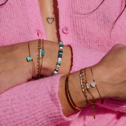 Superposition tendance de bracelets délicats en or rose, perles et pierres (turquoise, blanches) sur fond de cardigan rose vif. Tatouage cœur.