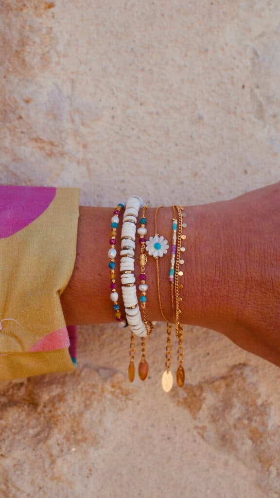 Bracelets Bohèmes : Bijoux Tendance avec Perles et Marguerite Bracelets bohèmes colorés avec perles et marguerite dorée sur un poignet devant un mur de pierre.